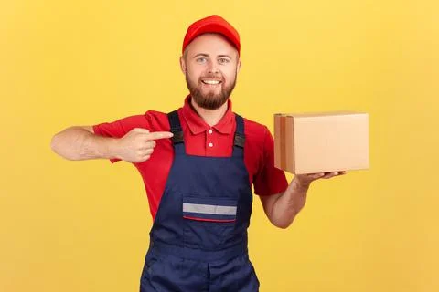 Friendly courier man in uniform pointing at cardboard box, delivering parcel to Stockfoto's