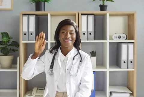 Friendly doctor looking at camera and waving hand having online video call or Stock Photos