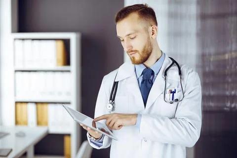 Friendly red-bearded doctor using tablet computer in clinic at his working place 库存照片