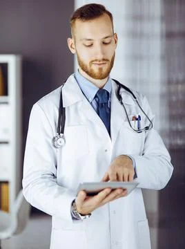 Friendly red-bearded doctor using tablet computer in clinic at his working place Photos