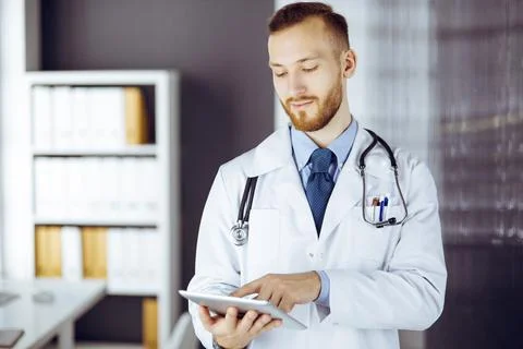 Friendly red-bearded doctor using tablet computer in clinic at his working place Stockfoto's