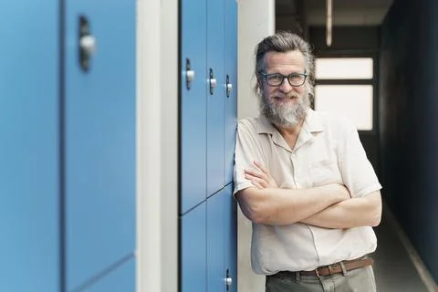 Friendly senior male teacher with beard standing in school hallway with blue Stock Photos