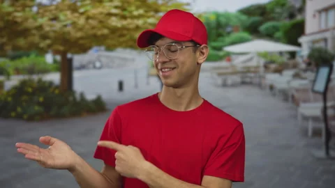 Friendly smiling man in red shirt and cap pointing finger to open hand at r.. Vidéo 315083215