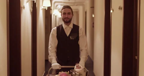 Friendly waiter in uniform rolling trolley in hotel corridor Stock Footage 233189052