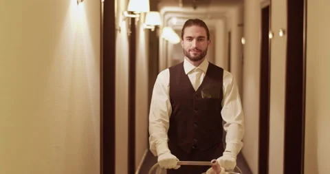 Friendly waiter in uniform rolling trolley in hotel corridor Stock Footage 233193386