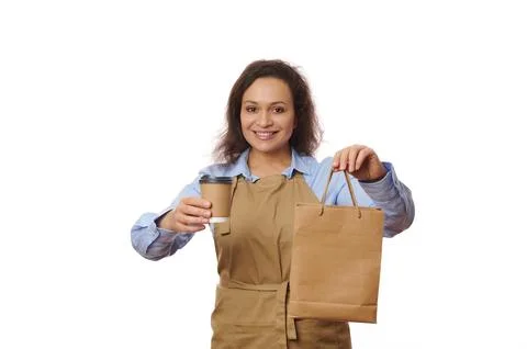 Friendly woman barista holds takeaway drink in paper cup, cutely smiling as Stock Photos