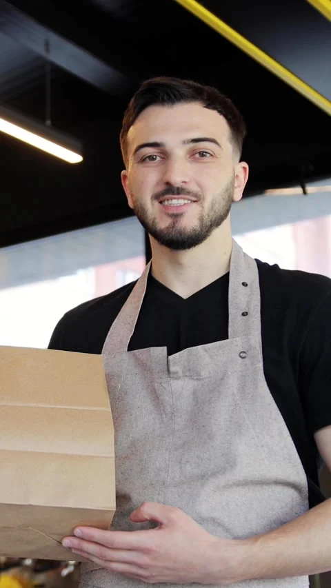Friendly young guy in apron holding paper package with take away meal and coffee Stock Footage 280386492