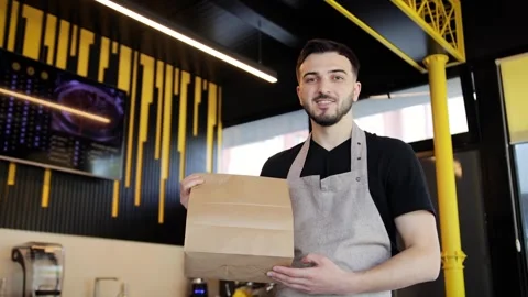 Friendly young man in apron holding paper package with take away meal and coffee Stock Footage 281248150