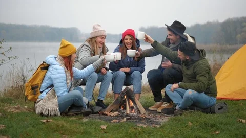Friends are clinking with drinks sitting around fire in forest with warm Stock Footage 119169357