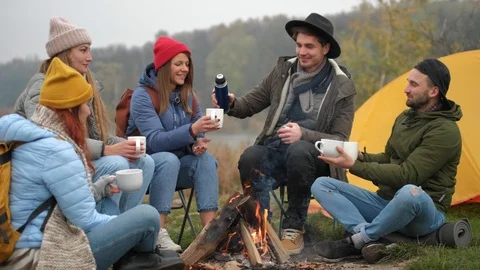 Friends are clinking with drinks sitting around fire in forest with warm Stock Footage 119169516