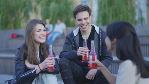 Friends are sitting on the steps, the guy is making a toast, they are drinking. Vidéo 202311736