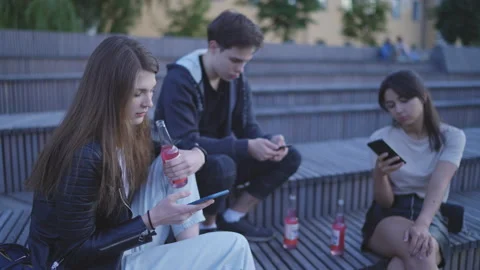 Friends are sitting on the steps, looking unemotionally at their phones. Summer. 库存影片 202313163
