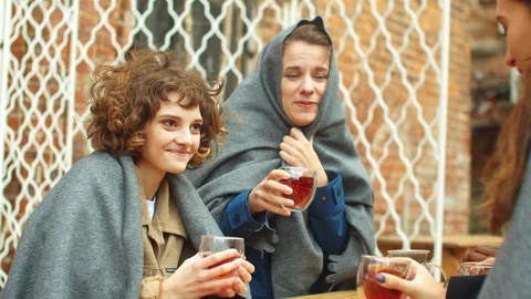 Friends in grey blankets sit outside the cafe, drink hot tea and talk Stock Footage 121941112