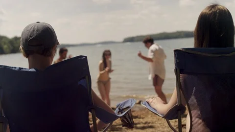 Friends sitting on chairs join their friends dancing on the beach at the lake Stock Footage