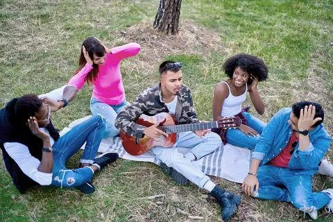Friends sitting on the ground while one plays a horrible guitar in a park. 스톡 사진