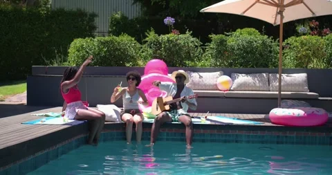 Friends sitting at pool edge, man starting strumming guitar, women lifting Stock Footage 335683698