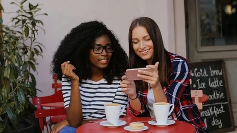 Friends sitting at table in cafe and using smartphone. Two girls looking at Stock Footage 81834140