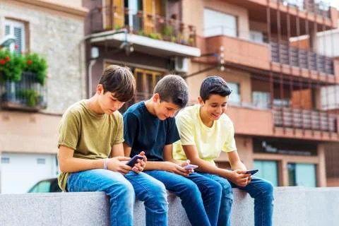 Friends using smart phones while sitting on retaining wall against building in Fotos de archivo