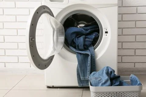 Front load washing machine with denim jeans inside and laundry basket Stock Photos