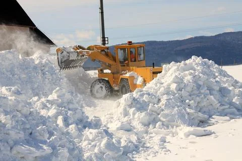 Front loader clears large piles of snow near rural buildings in winter, Stock Photos