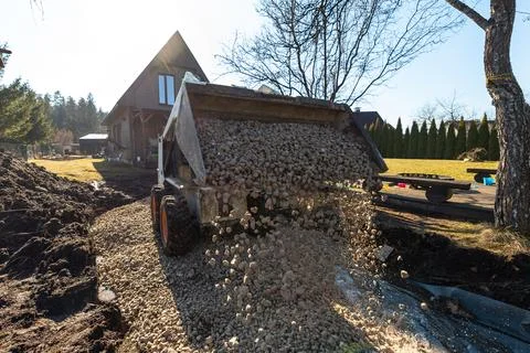 Front loader pours gravel onto path beside dark wood A frame cabin Stock Photos