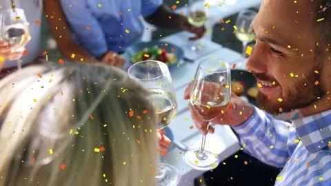 Front pair raising wine glasses, clinking then sipping at table, confetti Stockbeeldmateriaal 333602364