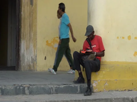 Front view Afro-American man dressed red t-shirt, trousers, hat, glasses sits Stock Footage 69558686