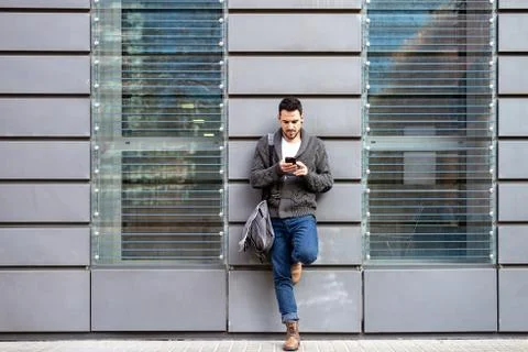 Front view of a bearded man using phone leaning on office building wall Fotos de archivo
