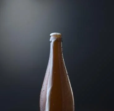 Front view of a bottle of cold beer with overflowing foam against dark. Stock Photos