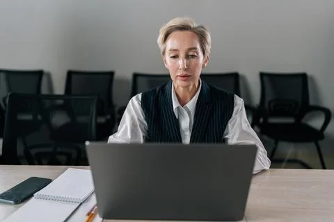 Front view of confident middle-aged business woman in suit sitting at desk Stock Photos