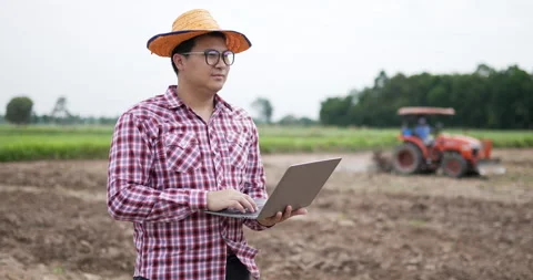 Front view of farmer standing and using laptop at farm with tractor background 스톡 동영상 200929831