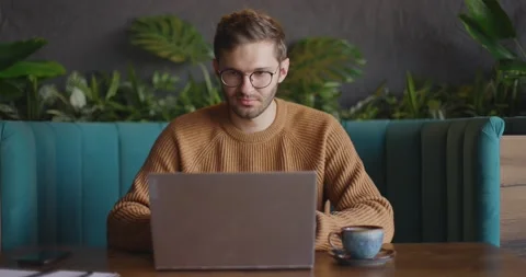 Front view of handsome man in eyeglasses in brown sweater finishes work at Stock Footage 164927113