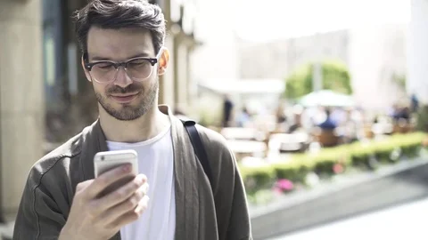 Front view of a hipster guy in glasses texting and smiling outside 스톡 동영상 77283648