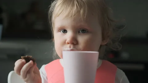 Front view of little cute white girl blonde with pink bib drinks kefir from pale Video stock 148380136