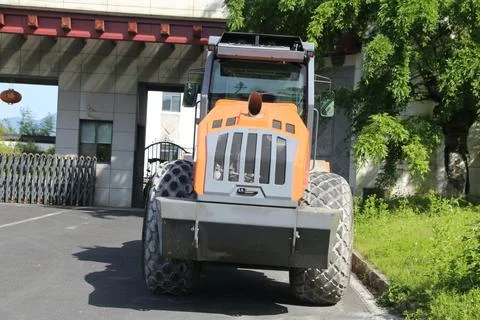 Front view of an orange road roller parked on fresh asphalt beside trees an.. Stock Photos