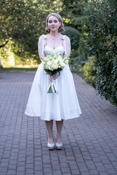 Front view of a serene bride in a tea-length white wedding dress, gently holding Stock Photos