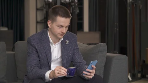 A front view of a young man sitting on sofa with the phone while having a cup of Stock Footage 108430755