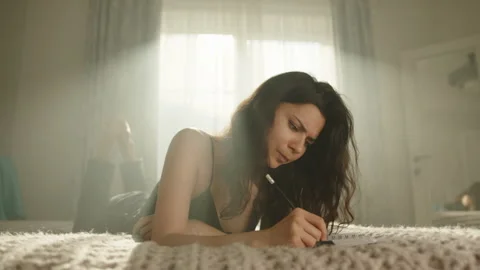 Frowning cute brunette making notes in notebook, lying on stomach on bed Stock Footage 244377609