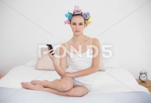 Frowning natural brown haired woman in hair curlers holding a mobile phone Stock Photos