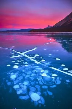 Frozen Lake Ice Bubbles at Sunset with Mountains and Vibrant Winter Sky Stock Photos