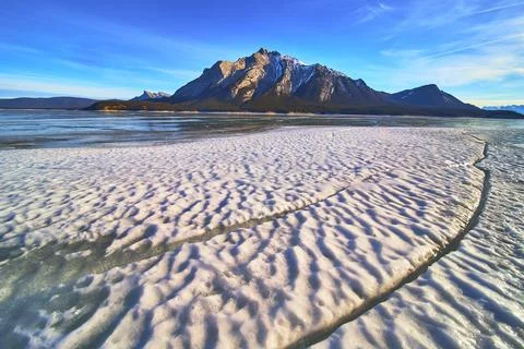 Frozen Lake Ice Patterns and Snowy Mountain Peaks Under Blue Winter Sky 스톡 사진