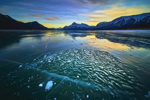 Frozen Lake Ice Patterns and Snow Capped Mountains at Vibrant Winter Sunset Stock Photos