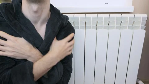 Frozen man in bathrobe shivering next to white heating radiator Stock Footage 328277455