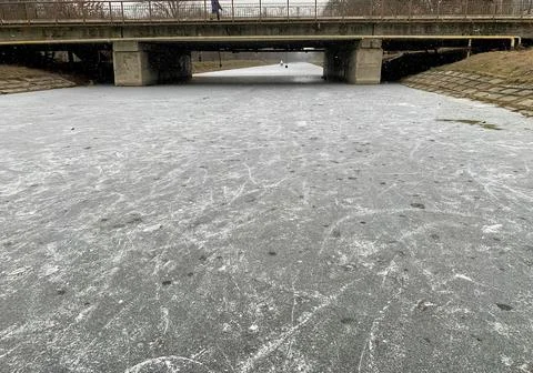 A frozen river flows beneath a concrete bridge in a city park during winter. The Stock Photos