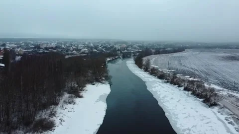 Frozen river flows near small town during cold winter day with snow covering lan Stock Footage 329708363