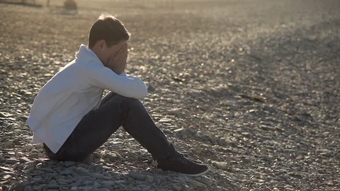 A frustrated 16-year- old boy in a white jacket sits on the beach in the wind, c Vidéo 114159115