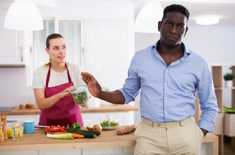 Frustrated man refusing container of vegetable salad offered by wife in kitchen Stock Photos