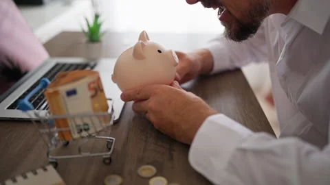 Frustrated man shaking an empty piggy bank at his desk, highlighting the co.. Stock Footage 340144179