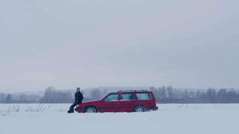 Frustrated man sit on hood of red broken down car in white winter snow landscape Stock Footage 332136728