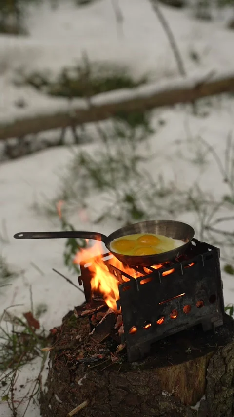 Frying eggs in a pan on a portable camping stove with an open flame, set on a 库存影片 313292563